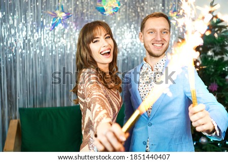 Brightfull expressions of happy emotions, laughing and celebrating together, couple having fun with sparklers over silver shiny background.