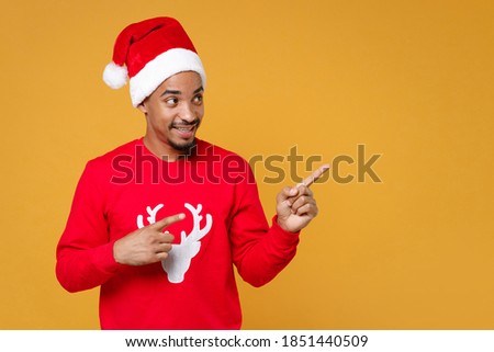 Smiling Santa african man in sweater Christmas hat pointing fingers aside on mockup copy space isolated on yellow background studio. Happy New Year celebration holiday concept. Tattoo translation life
