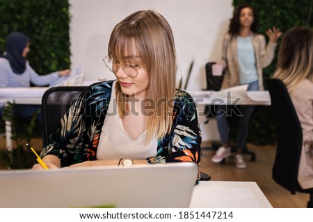 Portrait of a young female businesswoman working in an office while sitting at a table with colleagues in the background.