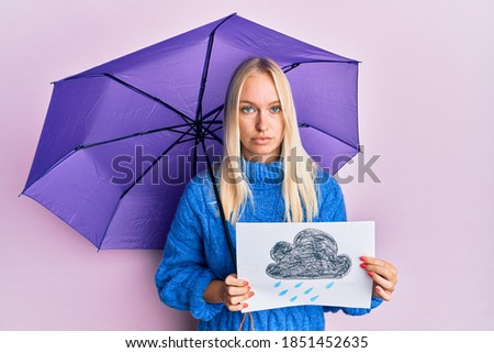 Young blonde girl holding umbrella rain draw relaxed with serious expression on face. simple and natural looking at the camera. 
