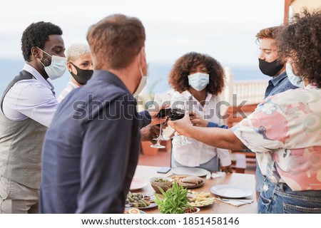 Young people enjoy dinner at home on patio and cheering with wine while wearing protective face mask for coronavirus - Focus on the hands