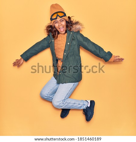 Young beautiful latin woman wearing winter clothes smiling happy. Jumping with smile on face and arms opened over isolated yellow background