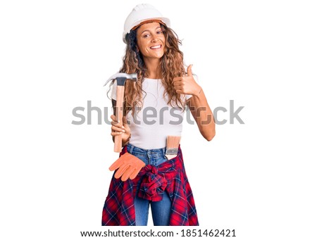 Young hispanic woman with tattoo wearing hardhat and builder clothes holding hammer smiling happy and positive, thumb up doing excellent and approval sign 