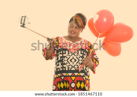 Happy fat black African woman smiling while holding bunch of red balloons with heart sign and taking selfie picture with selfie stick