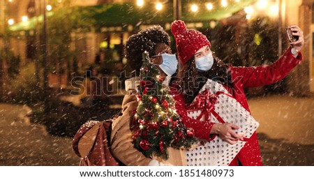 Portrait of joyful mixed-races females in medical masks standing on decorated street with x-mas presents and taking selfie photos on smartphone while snowing. Happy women taking pictures on cellphone