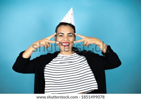 Young beautiful woman wearing a birthday hat over insolated blue background Doing peace symbol with fingers over face, smiling cheerful showing victory