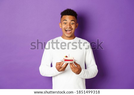 Image of happy african-american guy celebrating birthday, looking excited at b-day cake and making wish, standing over purple background