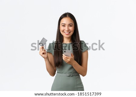 Small business owners, women entrepreneurs concept. Smiling attractive asian woman in dress making online purchase, buying tickets for work flight, holding mobile phone and credit card pleased