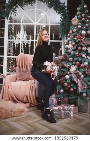 A beautiful blonde girl in a black sweater sits near a New Year tree with a gift in her hands. A gift for the new year in the hands of a girl. New Year mood