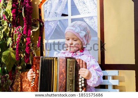 A little girl with a musical instrument accordion sitting at the window.