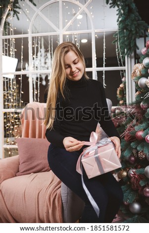 A beautiful blonde girl in a black sweater sits near a New Year tree with a gift in her hands. A gift for the new year in the hands of a girl. New Year mood