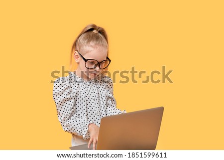 Cute baby girl in a shirt looks at an open laptop. Large glasses for vision. Yellow isolated background.