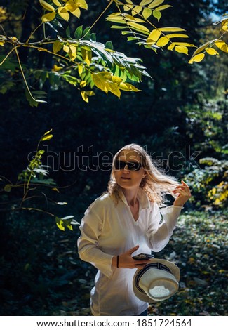 Cute blonde in white shirt with hat in her hand in the sunlight enjoying a walk in the park on beautiful autumn day. Young woman touches her hair and smiles.