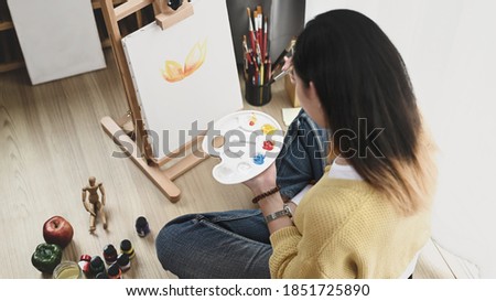 A young female artist with a palette in her hand painting on canvas while sitting on floor at her workshop.