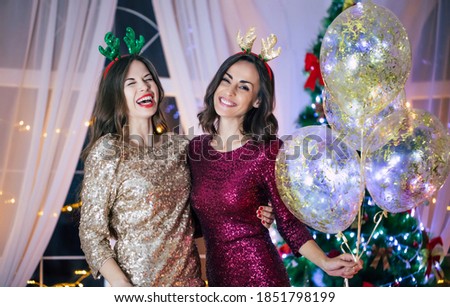 Two excited beautiful smiling girl friends in evening dresses are posing on the christmas tree background and having fun at the new year party