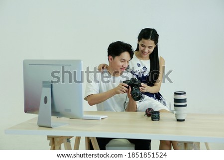 Young Asian couple photographer and model sitting in front of computer and desk, looking to screen camera while working process picture after shooting photo. Studio Photography Relation Hobby Concept