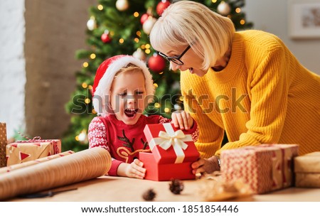 Excited kid in Santa hat opening gift box from granny while celebrating Christmas holiday at home
