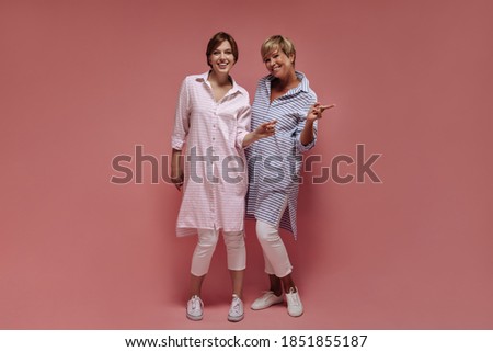 Positive two ladies with short cool hairstyle in modern striped shirt and skinny white pants smiling and showing peace signs on pink backdrop..