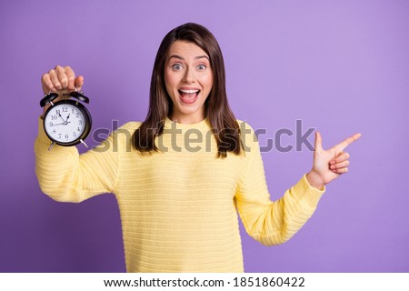 Photo portrait of positive woman holding clock with one hand pointing finger to side with open mouth isolated on vivid purple colored background