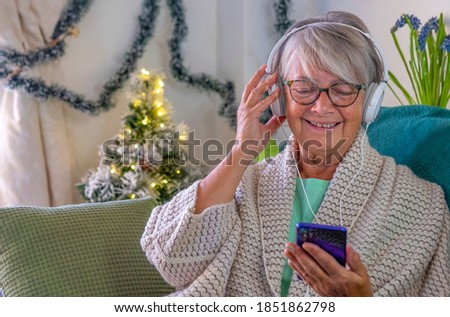 Attractive senior woman sitting at home on the sofa while listening to music with headphones while holding her smart phone. Christmas decoration on the background