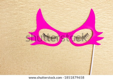 Paper accessory on a stick for a photo shoot for holidays or parties. Pink paper mask and false eyelashes on a gold background.