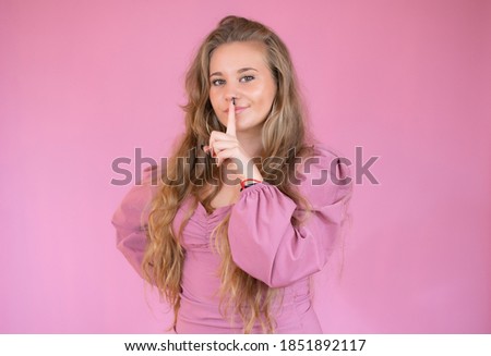 Pretty young female with cheerful expression, tells her secret to close friend, makes silence sign, stands against pink background with copy space aside. People, privacy and secrecy concept.