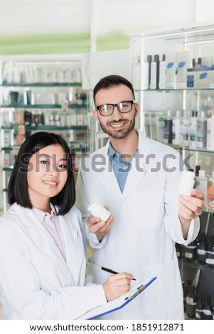 smiling pharmacist in white coat holding bottles with medication near asian colleague in drugstore