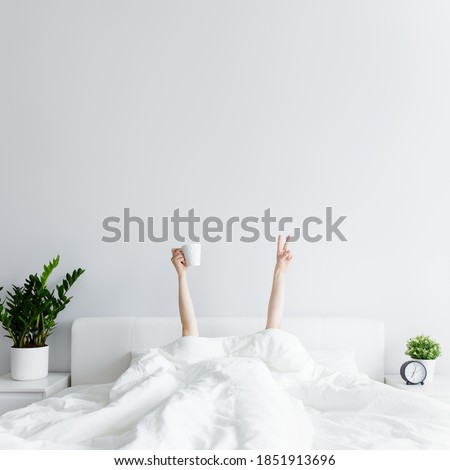 good morning concept - female hands with coffee cup and victory sign sticking out from the blanket at home or hotel, copy space over white wall background Royalty-Free Stock Photo #1851913696