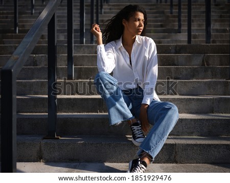 Waiting. Full length of beautiful young mixed race woman in casual wear looking thoughtful aside while sitting on stairs outdoors on a warm sunny day. Selective focus. Medium format shot