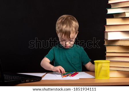 A small child sits at a table with books, a laptop and chooses a colored pencil. distance learning during quarantine. boy is doing homework. European appearance.