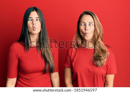 Hispanic family of mother and daughter wearing casual clothes over red background making fish face with lips, crazy and comical gesture. funny expression. 