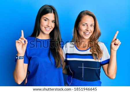 Hispanic family of mother and daughter wearing casual clothes over blue background with a big smile on face, pointing with hand finger to the side looking at the camera. 