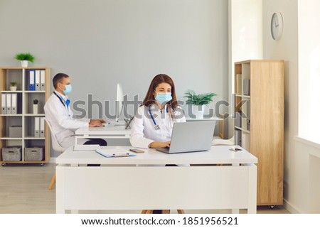 Doctors wearing medical face covering masks working on computers sitting at desks in hospital office. Healthcare workers and clinic staff care about colleagues and patients helping prevent coronavirus