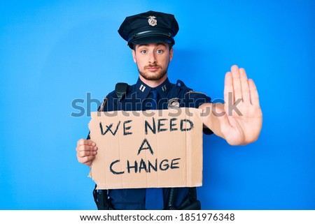 Young caucasian man wearing police uniform holding we need a change banner with open hand doing stop sign with serious and confident expression, defense gesture 