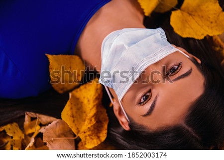 Beautiful young woman wearing a  medical mask and with a yellow maple leaf in hand covering her face. The scenery of the autumn park on a sunny day. Autumn season concept. Close up portrait.