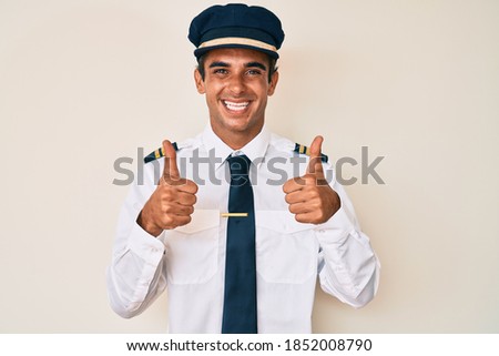 Young hispanic man wearing airplane pilot uniform success sign doing positive gesture with hand, thumbs up smiling and happy. cheerful expression and winner gesture. 