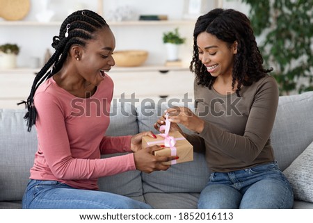 Excited black woman opening birthday gift from her girlfriend, living room interior, free space. Happy african american besties celebrating birthday at home, having fun, opening present