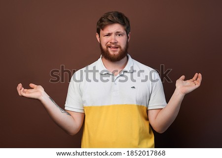 man throws up his hands while standing on brown isolate. disgruntled, frustrated bearded guy in t-shirt