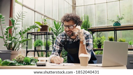 Happy Caucasian businessman talking on cellphone while standing in apron in small floral center and writing down order details. Joyful male florist calling on smartphone at work. Own business concept Royalty-Free Stock Photo #1852040431