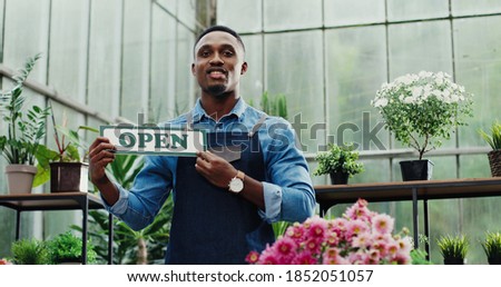 Portrait of young African American male employee standing at floral shop and holding Open sign in hands and smiling to camera. Businessman opens store after quarantine. Retail concept