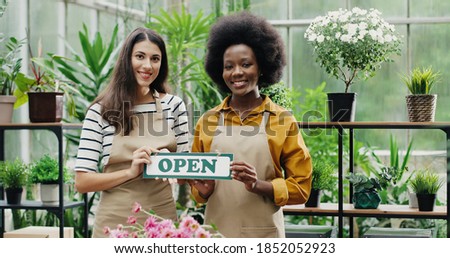 Happy mixed-races women florists in aprons standing in own floral shop and smiling to camera with Open sign in hands. Caucasian and African American females reopening flower store. Portrait concept