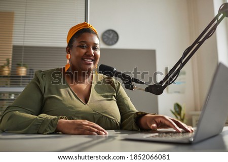 Portrait of African happy woman smiling at camera while sitting at the table with laptop and leading live broadcast