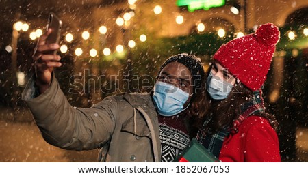 Close up of joyful African American man with Caucasian woman on street while taking selfie photo on smartphone. Happy multi-ethnic couple standing outdoors and taking pictures together with xmas gift