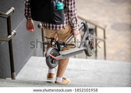 Man Carrying folded E-Scooter in hand on stairs near building. Moving by eco urban transport, modern city concept. Ecological technological lifestyle. Person Carry Electric Scooter In Folded Position. Royalty-Free Stock Photo #1852101958