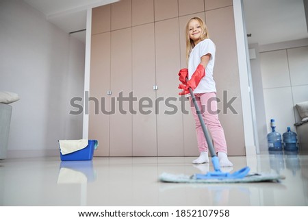 Low angle portrait of happy smiling little girl with floor mop in hand while posing at the photo camera in apartments