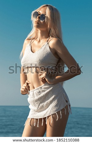 A girl with blond hair is sunbathing while standing by the turquoise sea on a clear sunny day in sunglasses and in a light knitted swimsuit