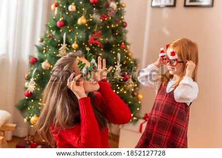 Mother and daughter having fun playing at home on Christmas day, sitting near nicely decorated Christmas tree and trying on party glasses