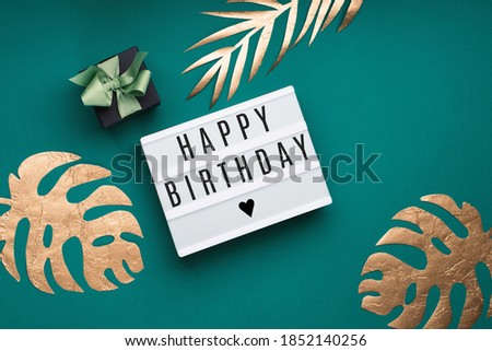Happy Birthday text on white Lightbox, gift box and golden tropical leaves Monstera on paper blue background. Festive greeting card concept. Flat lay, top view, copy space.
