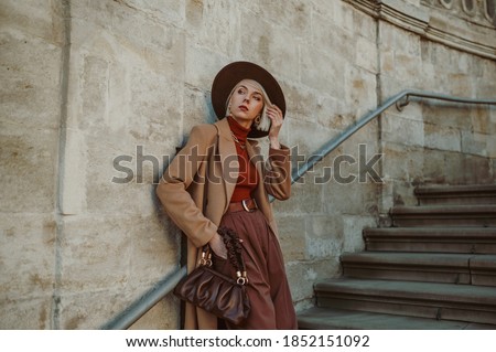 Outdoor autumn fashion portrait of elegant, luxury woman wearing trendy midi beige, camel color coat, brown hat, trousers, orange turtleneck, holding leather pouch bag, posing in street of city Royalty-Free Stock Photo #1852151092