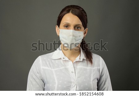 Young woman in a medical mask and gown, standing and looking straight into the camera, portrait, head slightly tilted to the side.